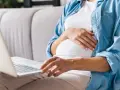 Kobieta w ciąży siedzi na kanapie, pisząc na laptopie wniosek o urlop macierzyński.
