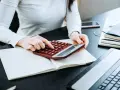 Kobieta liczy wydatki na własne cele mieszkaniowe, przygotowując PIT-39. Na biurku notatnik, kalkulator i laptop.