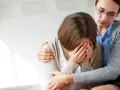 Kobieta płacze, ukrywając twarz w dłoniach. Druga kobieta obejmuje ją ramieniem, oferując wsparcie w trudnym czasie, np. po utracie ojca i staraniach o rentę po zmarłym ojcu.