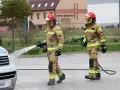 Dwóch strażaków w akcji. Jeden z nich polewa wodą samochód, drugi niesie wąż. Ciekawe, ile zarabia strażak za taką pracę.