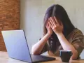 Kobieta z dłońmi na twarzy, przy laptopie, z kubkiem kawy. Objawy wypalenia zawodowego, czas na L4.