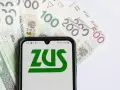 ZUS na ekranie telefonu, obok banknoty 100, 200 i 500 zł. Symbolizuje to zasiłek pogrzebowy 6000 zł.
