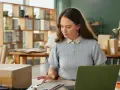 Kobieta pracuje przy biurku, na którym leży tablet z danymi. W tle widać regały z paczkami. Może to być związane z ksef odroczenie.