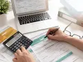 Ręce wypełniające formularz PIT-37, obok kalkulatora z wynikiem 236.12, laptopa i okularów.