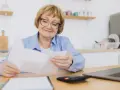 Uśmiechnięta starsza pani z okularami przegląda dokumenty, być może związane z rentą socjalną z MOPS.