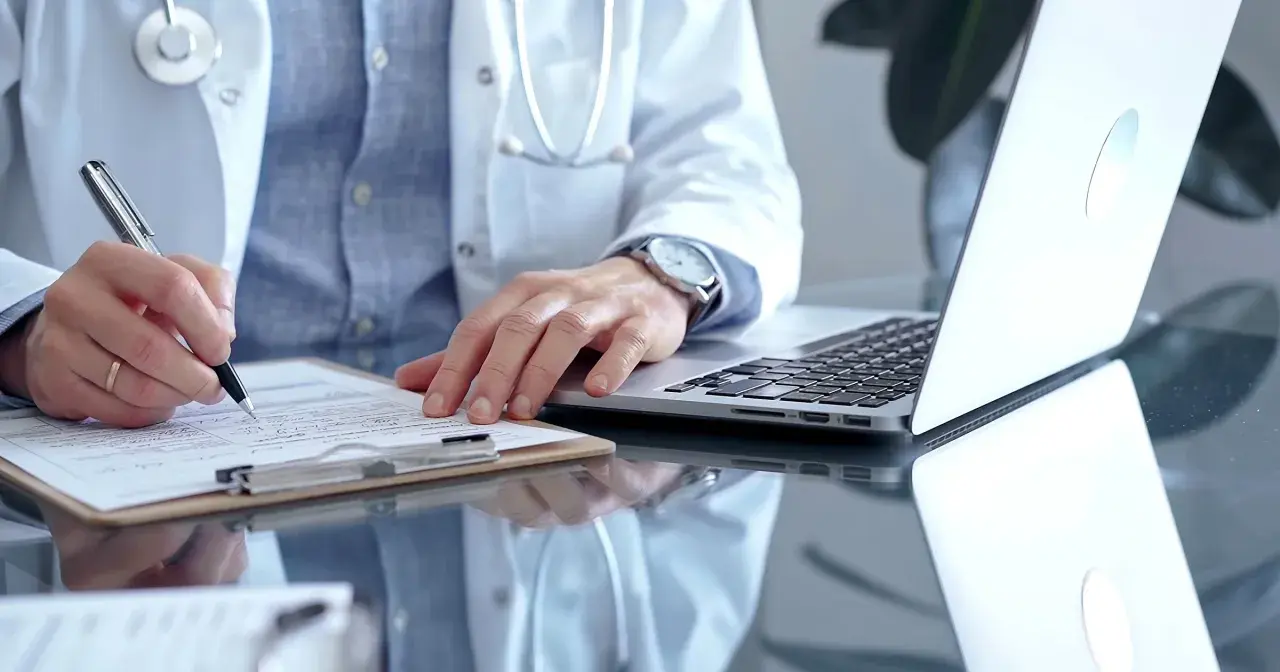 Lekarz w białym kitlu pisze na dokumentach, obok laptop. Szybka konsultacja psychiatryczna online.