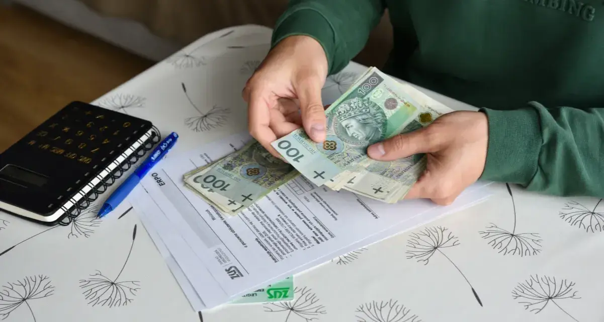 Ręce liczą banknoty 100 zł nad dokumentami ZUS. Obok leży kalkulator i długopis. To może być pomocne przy ustalaniu, ile wynosi świadczenie przedemerytalne na rękę.