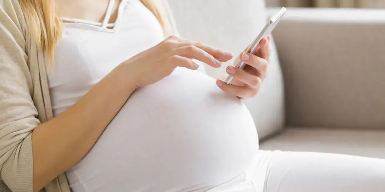 Kobieta w 34. tygodniu ciąży przegląda coś na telefonie, siedząc na kanapie.
