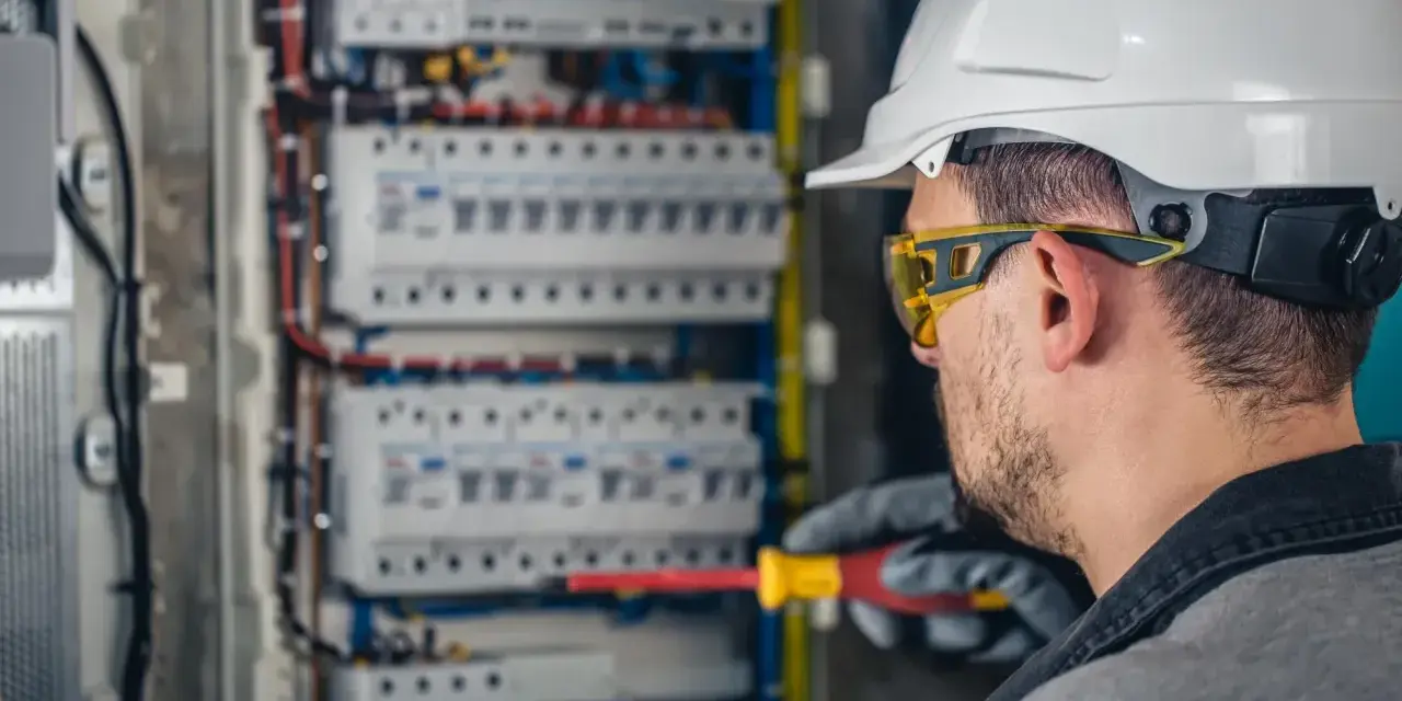 Elektryk w kasku i okularach ochronnych pracuje przy rozdzielnicy. Zastanawiasz się, ile zarabia elektryk?
