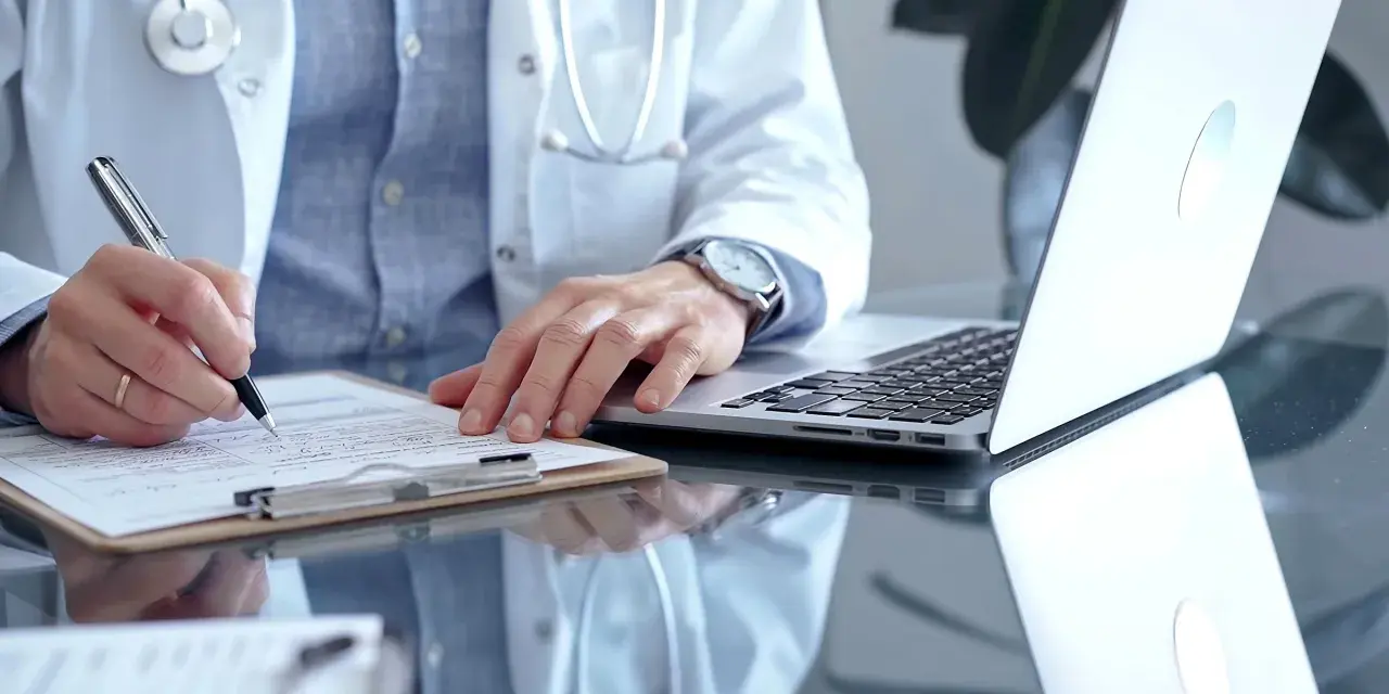 Lekarz w białym kitlu pisze na dokumentach, obok laptop. Szybka konsultacja psychiatryczna online.