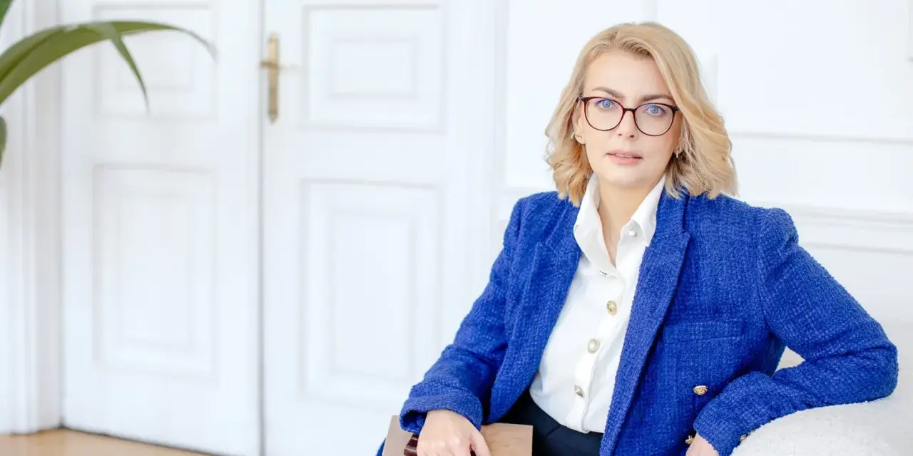Kobieta w okularach i niebieskiej marynarce trzyma notatnik. Planuje dofinansowanie na otwarcie firmy.