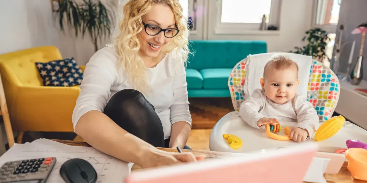 Uśmiechnięta mama pracuje przy laptopie, podczas gdy dziecko bawi się w krzesełku. Planuje zwrot podatku na dziecko.