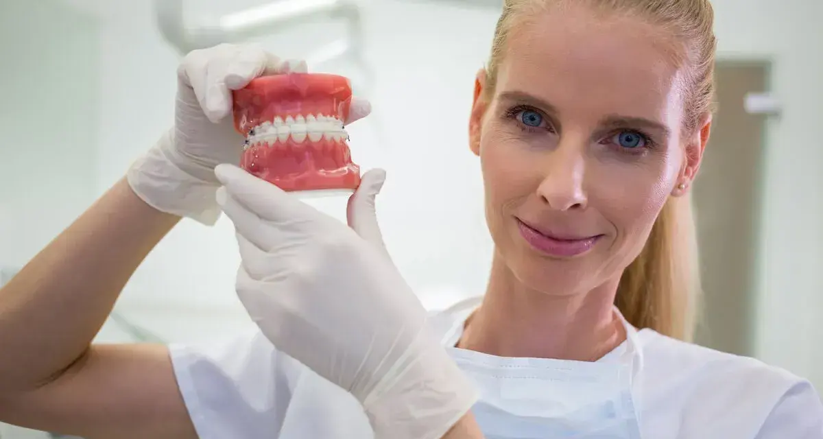 Dentystka w białej rękawiczce pokazuje model szczęki z aparatem ortodontycznym. Zastanawiasz się, ile zarabia dentysta?