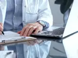 Lekarz w białym kitlu pisze na dokumentach, obok laptop. Szybka konsultacja psychiatryczna online.