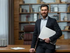 Uśmiechnięty dyrektor szkoły w garniturze, trzymający laptopa, stoi przy biurku w gabinecie.