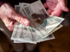Dłonie trzymają polskie banknoty 100 zł i monety. Ile trzeba przepracować żeby dostać emeryturę?
