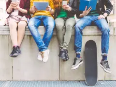 Cztery osoby siedzą na murku, każda zajęta swoim urządzeniem lub książką. To pokolenie z od kiedy, które ceni technologię i wiedzę. Obok leży deskorolka.