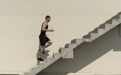 Mężczyzna w czarnym stroju sportowym wspina się po betonowych schodach na tle jasnej ściany.
