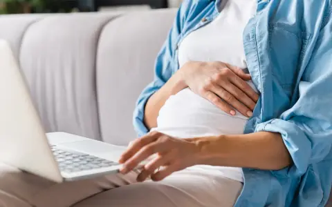 Kobieta w ciąży siedzi na kanapie, pisząc na laptopie wniosek o urlop macierzyński.