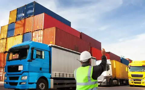 Pracownik w kamizelce kieruje ruchem ciężarówek z kontenerami. To właśnie spedycja co to – organizacja transportu.