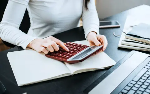Kobieta liczy wydatki na własne cele mieszkaniowe, przygotowując PIT-39. Na biurku notatnik, kalkulator i laptop.