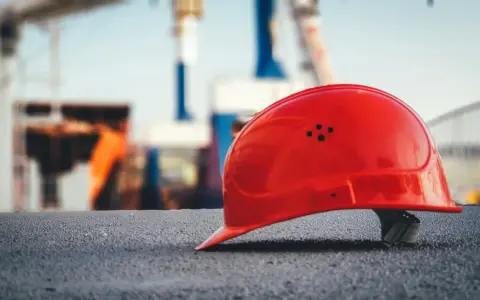 Czerwony kask budowlany na tle placu budowy. Symbol bezpieczeństwa pracy, który może wpływać na stopę procentową składek na ubezpieczenie wypadkowe.
