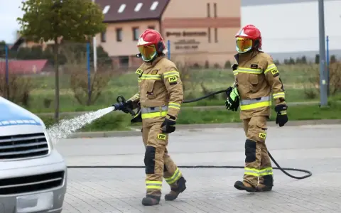 Dwóch strażaków w akcji. Jeden z nich polewa wodą samochód, drugi niesie wąż. Ciekawe, ile zarabia strażak za taką pracę.