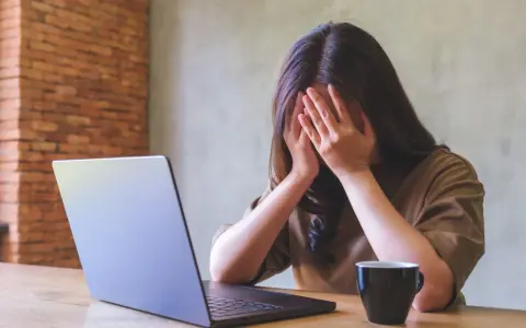 Kobieta z dłońmi na twarzy, przy laptopie, z kubkiem kawy. Objawy wypalenia zawodowego, czas na L4.
