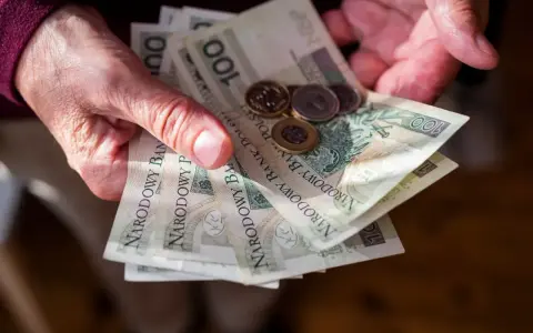 Dłonie trzymają polskie banknoty 100 zł i monety. Ile trzeba przepracować żeby dostać emeryturę?