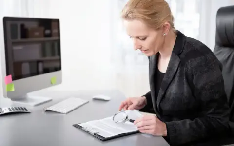 Pracownik samorządowy z lupą analizuje dokumenty. Na biurku komputer i klawiatura.