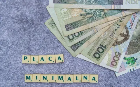 Płaca minimalna w Polsce – polskie banknoty o nominałach 100 i 500 zł ułożone na szarym tle, obok kafelki z literami tworzące napis "PŁACA MINIMALNA".