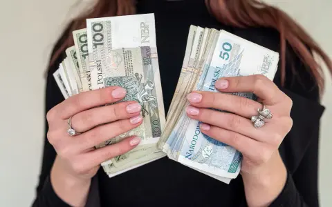 Dłonie z paznokciami w kolorze nude trzymają stosy banknotów 100 i 50 zł. To idealna jubileuszówka na prezent!