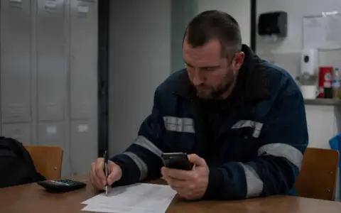 Pracownik w roboczym ubraniu sprawdza telefon, notując coś na papierze. Może to być jego dodatek nocny 2025.