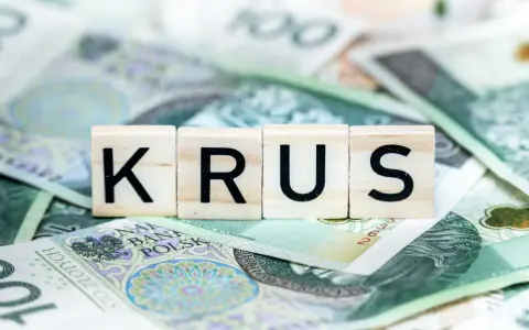 Drewniane klocki z napisem "KRUS" leżą na stosie polskich banknotów stuzłotowych. To symbol oszczędności na emeryturę z KRUS.