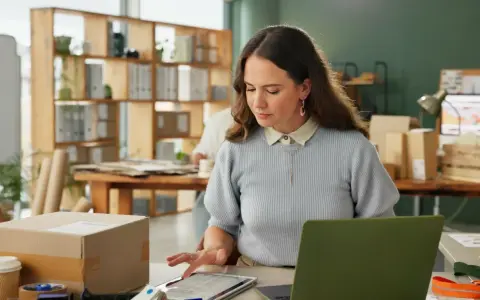 Kobieta pracuje przy biurku, na którym leży tablet z danymi. W tle widać regały z paczkami. Może to być związane z ksef odroczenie.