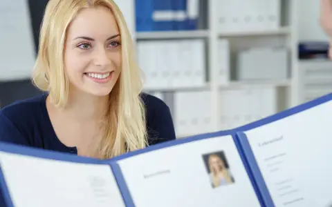 Uśmiechnięta blondynka prezentuje dokumenty, być może list motywacyjny wzór, podczas rozmowy kwalifikacyjnej.