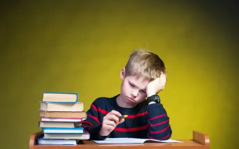 Chłopiec z podpartą głową siedzi przy biurku ze stosem książek i zeszytem.
