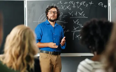 Nauczyciel w niebieskiej koszuli tłumaczy matematykę przy tablicy. Czy nauczyciel jest funkcjonariuszem publicznym? To pytanie nurtuje wielu.