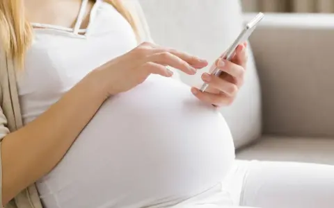 Kobieta w 34. tygodniu ciąży przegląda coś na telefonie, siedząc na kanapie.