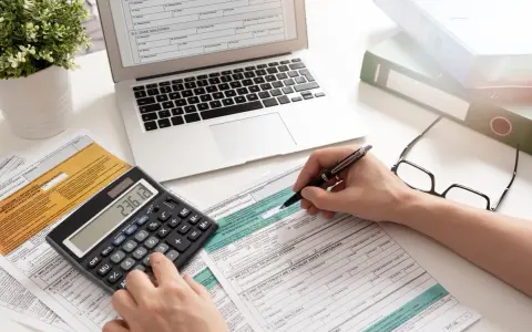 Ręce wypełniające formularz PIT-37, obok kalkulatora z wynikiem 236.12, laptopa i okularów.
