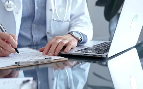 Lekarz w białym kitlu pisze na dokumentach, obok laptop. Szybka konsultacja psychiatryczna online.