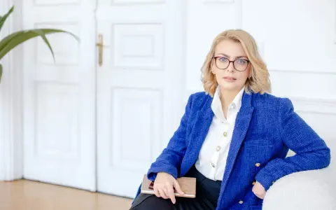Kobieta w okularach i niebieskiej marynarce trzyma notatnik. Planuje dofinansowanie na otwarcie firmy.