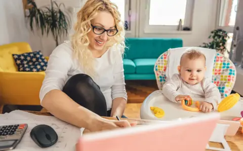 Uśmiechnięta mama pracuje przy laptopie, podczas gdy dziecko bawi się w krzesełku. Planuje zwrot podatku na dziecko.
