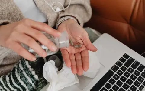 Dłoń z kapsułkami, obok laptop i chusteczki. Kto płaci za L4, gdy choroba dopada?