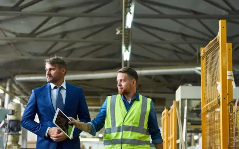 Mężczyzna w garniturze i pracownik w kamizelce odblaskowej omawiają stan bezpieczeństwa i higieny pracy w zakładzie, patrząc na tablet.