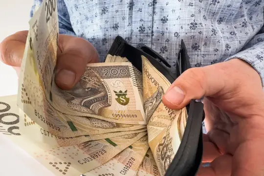 Dłonie trzymają portfel z banknotami 200 zł. Ile trzeba przepracować, by tyle zarobić?
