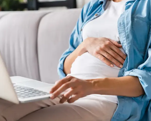 Kobieta w ciąży siedzi na kanapie, pisząc na laptopie wniosek o urlop macierzyński.