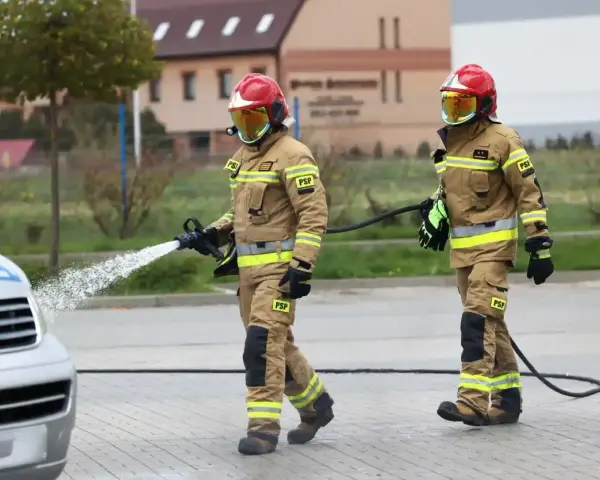 Dwóch strażaków w akcji. Jeden z nich polewa wodą samochód, drugi niesie wąż. Ciekawe, ile zarabia strażak za taką pracę.