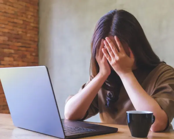 Kobieta z dłońmi na twarzy, przy laptopie, z kubkiem kawy. Objawy wypalenia zawodowego, czas na L4.