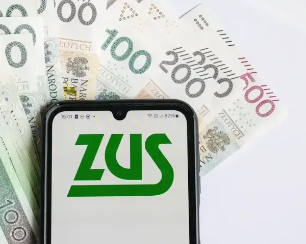 ZUS na ekranie telefonu, obok banknoty 100, 200 i 500 zł. Symbolizuje to zasiłek pogrzebowy 6000 zł.